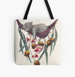 This is a tote bag with the image of a relaxing wombat in a hammock. The image is printed on both sides of the washable cloth bag. The hammock has gum leaves, nuts and flowers pinned to it. This image is painted by Patricia (PJ) Hopwood-Wade.